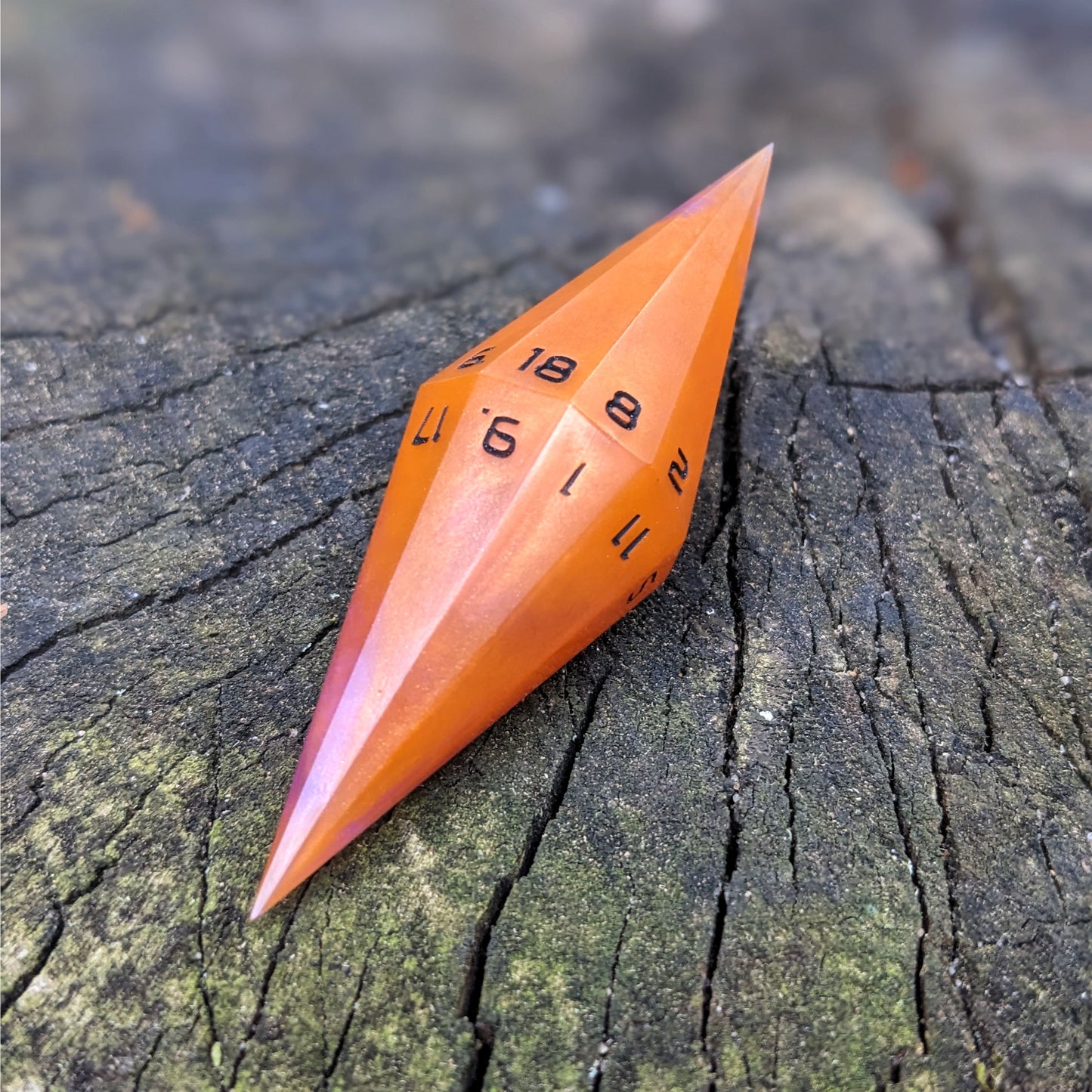 An orange and purple dagger shaped d20 with black numbers sits on a wood stump. The 18 face is on top.