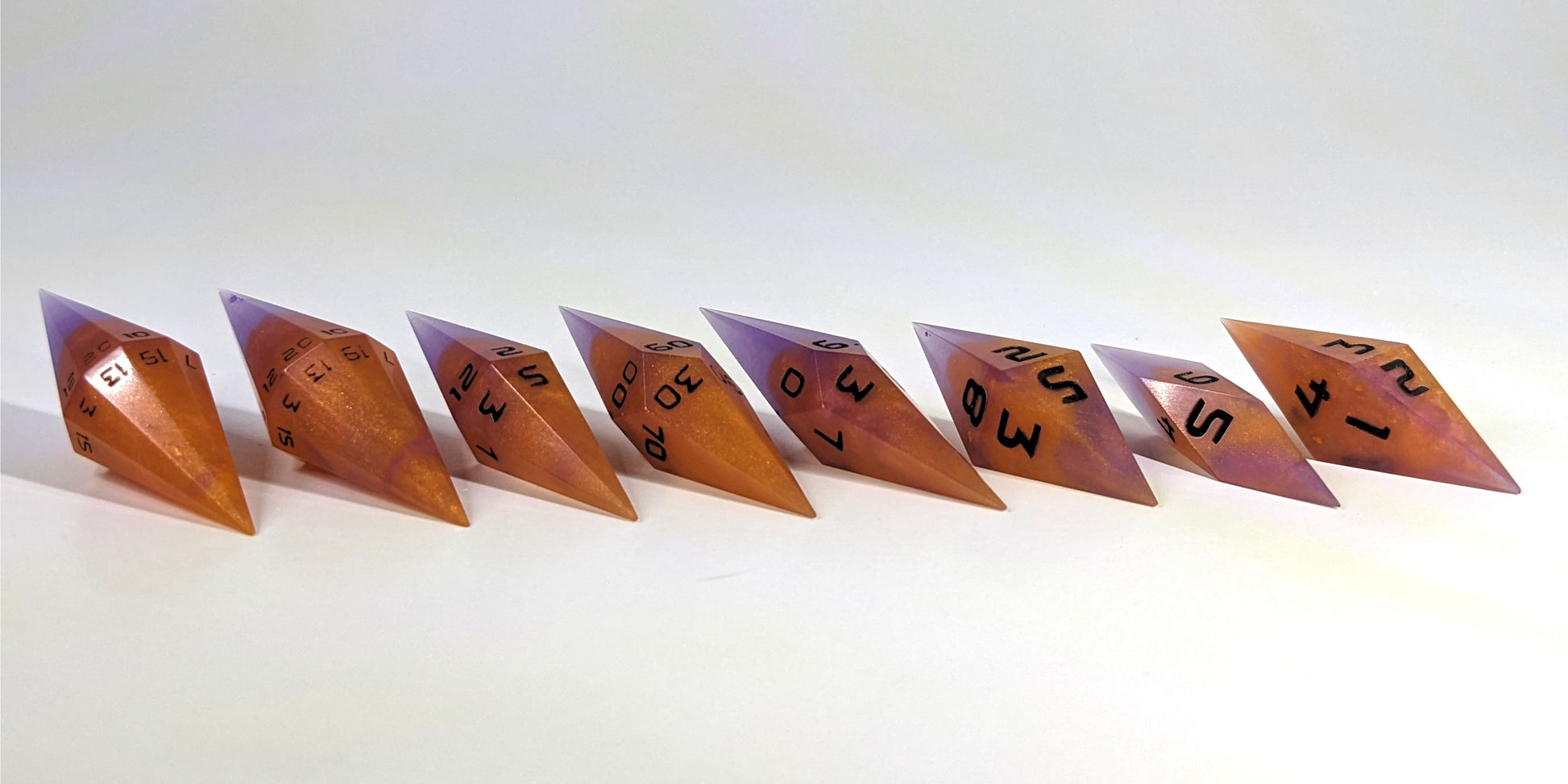 A straight on photo of a complete set of handmade diamond DnD dice that are purple and orange with black numbers arranged in a straight line with bottom points angled slightly forwards and towards the right. They are lined up with the 2 d20s on the left going down to the d4 on the right side. The highest value number of each die is facing forward.