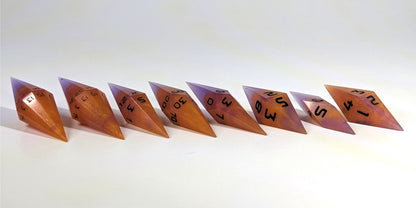 A straight on photo of a complete set of handmade diamond DnD dice that are purple and orange with black numbers arranged in a straight line with bottom points angled slightly forwards and towards the right. They are lined up with the 2 d20s on the left going down to the d4 on the right side. The highest value number of each die is facing forward.