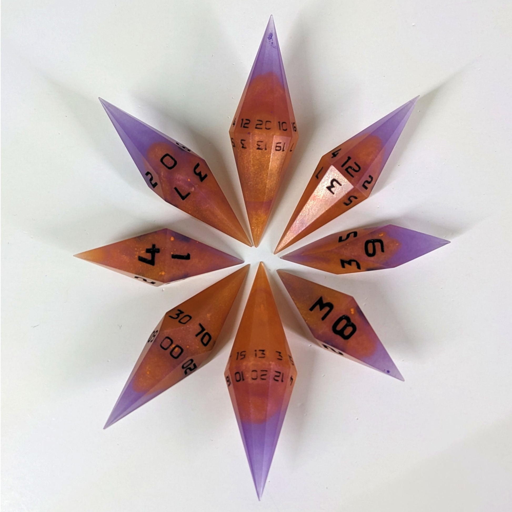 A set of handmade purple and orange diamond shaped dice with black numbers are arranged with the bottom points angled toward a center point, making the top points of the dice point outward in a circle. The highest value of each die is facing upwards, and the camera is shooting from straight above.