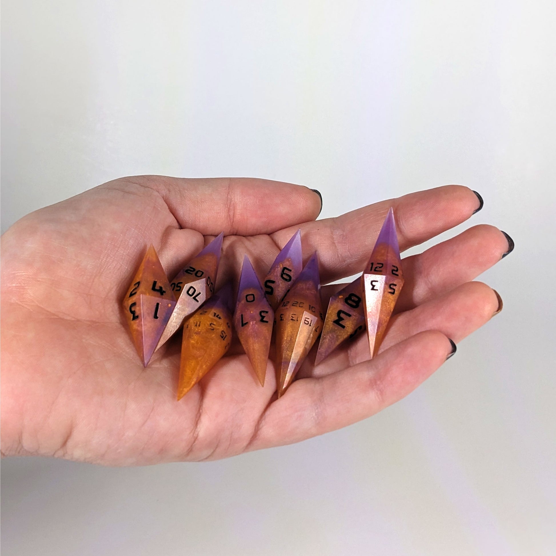 An 8 dice set of diamond style dice that are purple and orange with black numbers are held in a hand. The image shows that the set fits easily in a hand, making them overall smaller, though sharper, than a standard set of dnd dice.