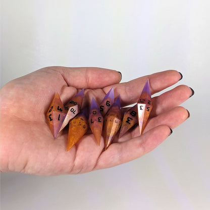 An 8 dice set of diamond style dice that are purple and orange with black numbers are held in a hand. The image shows that the set fits easily in a hand, making them overall smaller, though sharper, than a standard set of dnd dice.