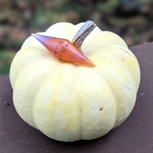 A diamond style d20 that is orange and purple with black numbers sits on top of a small, white pumpkin. The handmade die rests near the stem, and the 20 face is facing forward.