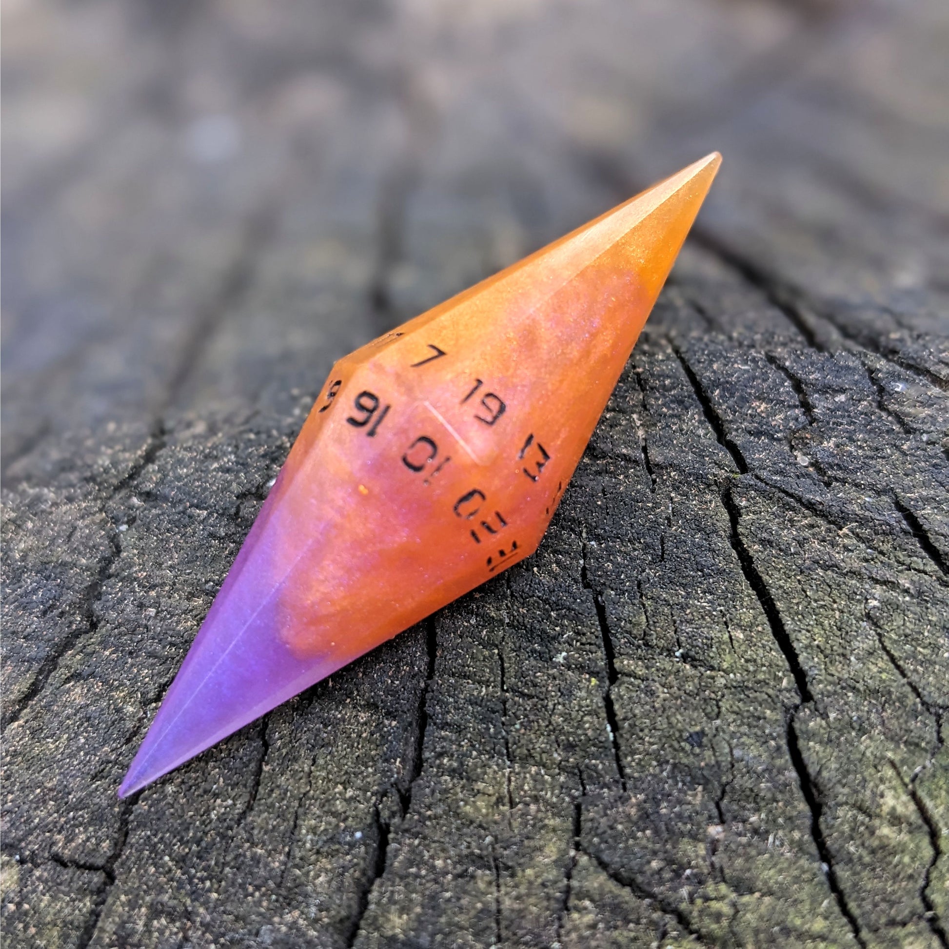 An orange and purple diamond shaped d20 with black numbers sits on a wood stump. The 7 face is on top.