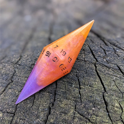 An orange and purple diamond shaped d20 with black numbers sits on a wood stump. The 7 face is on top.