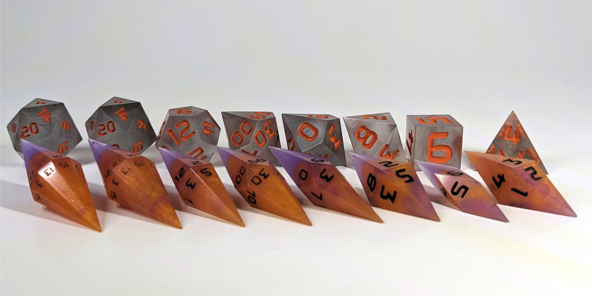 A straight on photo of a complete set of dagger dice that are purple and orange with black numbers arranged in a straight line with points angled forward and to the right. They are lined up with the 2 d20s on the left going down to the d4 on the right side. Behind the dagger set is a standard set of polyhedral dice to allow for size comparison. The diamond dice are much smaller overall. The highest value number of each die is facing forward.