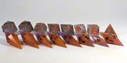 A straight on photo of a complete set of dagger dice that are purple and orange with black numbers arranged in a straight line with points angled forward and to the right. They are lined up with the 2 d20s on the left going down to the d4 on the right side. Behind the dagger set is a standard set of polyhedral dice to allow for size comparison. The diamond dice are much smaller overall. The highest value number of each die is facing forward.
