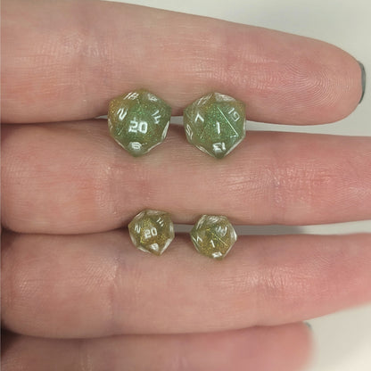 Two pairs of handmade d20 earrings with titanium hardware that are bright green and red gold with white numbers held between the fingers of a flat hand. The larger pair is between the pointer and middle finger, the smaller between the middle and ring finger. The earrings on the left of each pair centers the 20 face and the earrings on the right of each pair centers the 1 face.