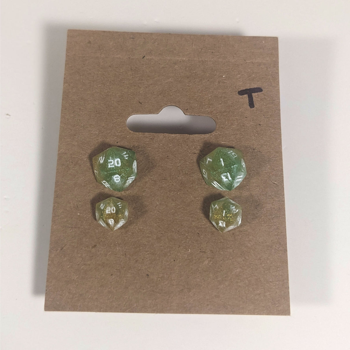 Two pairs of handmade d20 earrings with titanium hardware that are bright green and red gold with white numbers on a kraft brown earring card with a T in the top right corner. The earrings on the left of each pair centers the 20 face and the earrings on the right of each pair centers the 1 face.