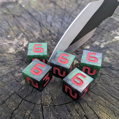 Five six sided dice sit on a wood stump with a throwing knife stabbed into the wood. The handmade dice are dark green and black with red numbers.