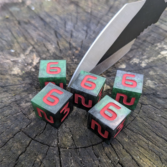 Five six sided dice sit on a wood stump with a throwing knife stabbed into the wood. The handmade dice are dark green and black with red numbers.