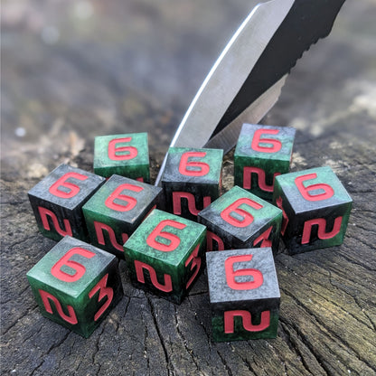 Ten six sided dice sit on a wood stump with a throwing knife stabbed into the wood. The handmade dice are dark green and black with red numbers.