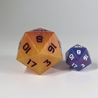 A straight on shot of a handcrafted large d20 that has swirls of orange, yellow, and a small amount of red with dark red numbers to the left of a standard size d20 that has swirls of blue, pink, and purple with some sparkle. This size comparison shows the large d20 being about twice the size of the standard.