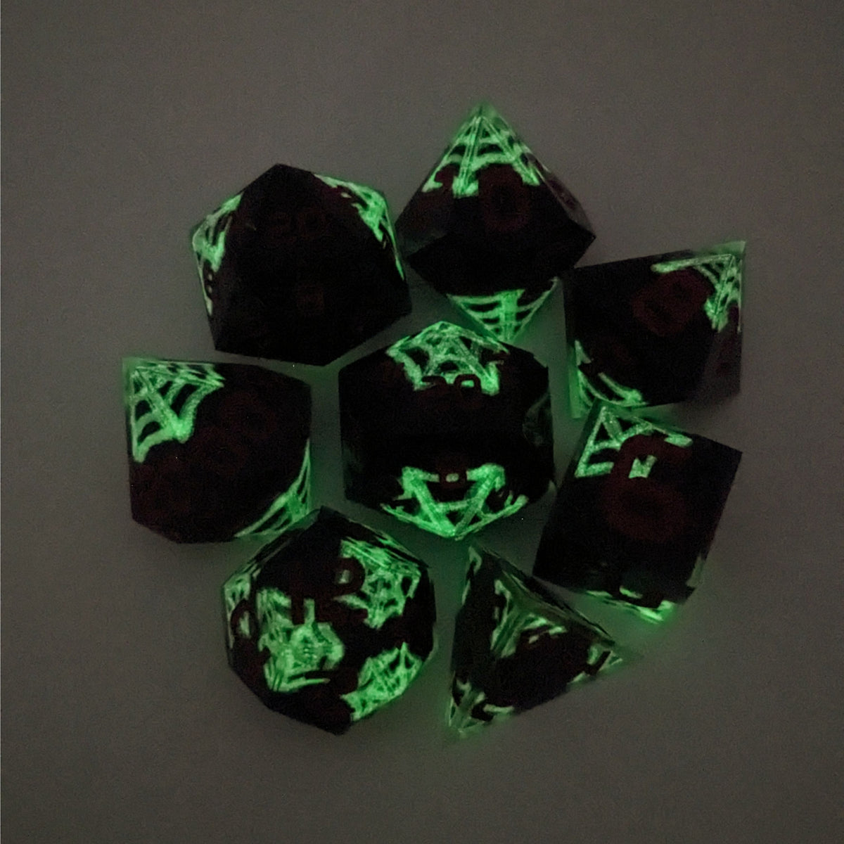 A complete set of 8 handmade artisan dice that are black with hand painted white spider webs that glow in the dark and red numbers. The DnD dice set is arranged in a circle and features 2 D20s, one of which is in the center surrounded by the other 7 dice on a white background. This photo is taken in the dark to show the white webs glowing green.