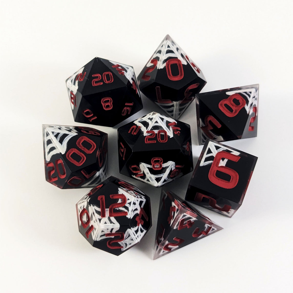 A complete set of 8 handmade artisan dice that are black with hand painted white spider webs that glow in the dark and red numbers. The DnD dice set is arranged in a circle and features 2 D20s, one of which is in the center surrounded by the other 7 dice on a white background.