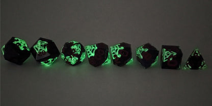 A straight on photo of a complete set of polyhedral dice that are black with hand painted white spider webs that glow in the dark and red numbers arranged in a straight line. They are lined up with the 2 d20s on the left going down to the d4 on the right side. The highest value number of each die is facing forward. This photo is taken in the dark to show the spider webs glowing green in the dark.