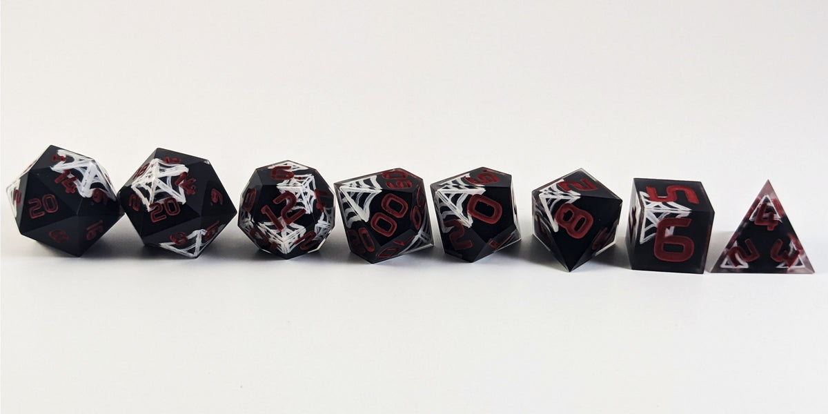 A straight on photo of a complete set of polyhedral dice that are black with hand painted white spider webs that glow in the dark and red numbers arranged in a straight line. They are lined up with the 2 d20s on the left going down to the d4 on the right side. The highest value number of each die is facing forward