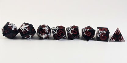 A straight on photo of a complete set of polyhedral dice that are black with hand painted white spider webs that glow in the dark and red numbers arranged in a straight line. They are lined up with the 2 d20s on the left going down to the d4 on the right side. The highest value number of each die is facing forward