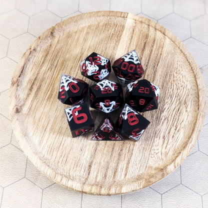 A complete set of 8 handmade artisan dice that are black with hand painted white spider webs that glow in the dark and red numbers. The DnD dice set is on a wooded circle and the dice are arranged in a circle and features 2 D20s, one of which is in the center surrounded by the other 7 dice.