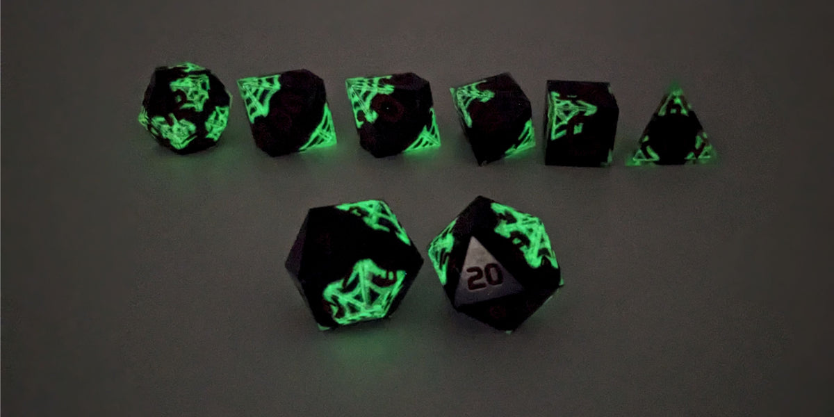 A straight on photo of a standard set of DnD dice that are black with hand painted white spider webs that glow in the dark and red numbers focusing on 2 d20s in the center. The d20 on the left has the 1 face centered and the d20 on the right has the 20 face centered. The other 6 dice are lined up in the background but are out of focus, while the d20s are in sharp focus. This photo is taken in the dark to show the webs glowing green in the dark.