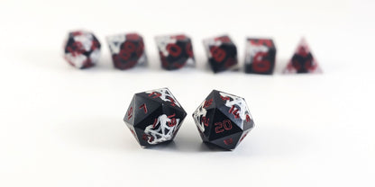 A straight on photo of a standard set of DnD dice that are black with hand painted white spider webs that glow in the dark and red numbers focusing on 2 d20s in the center. The d20 on the left has the 1 face centered and the d20 on the right has the 20 face centered. The other 6 dice are lined up in the background but are out of focus, while the d20s are in sharp focus.