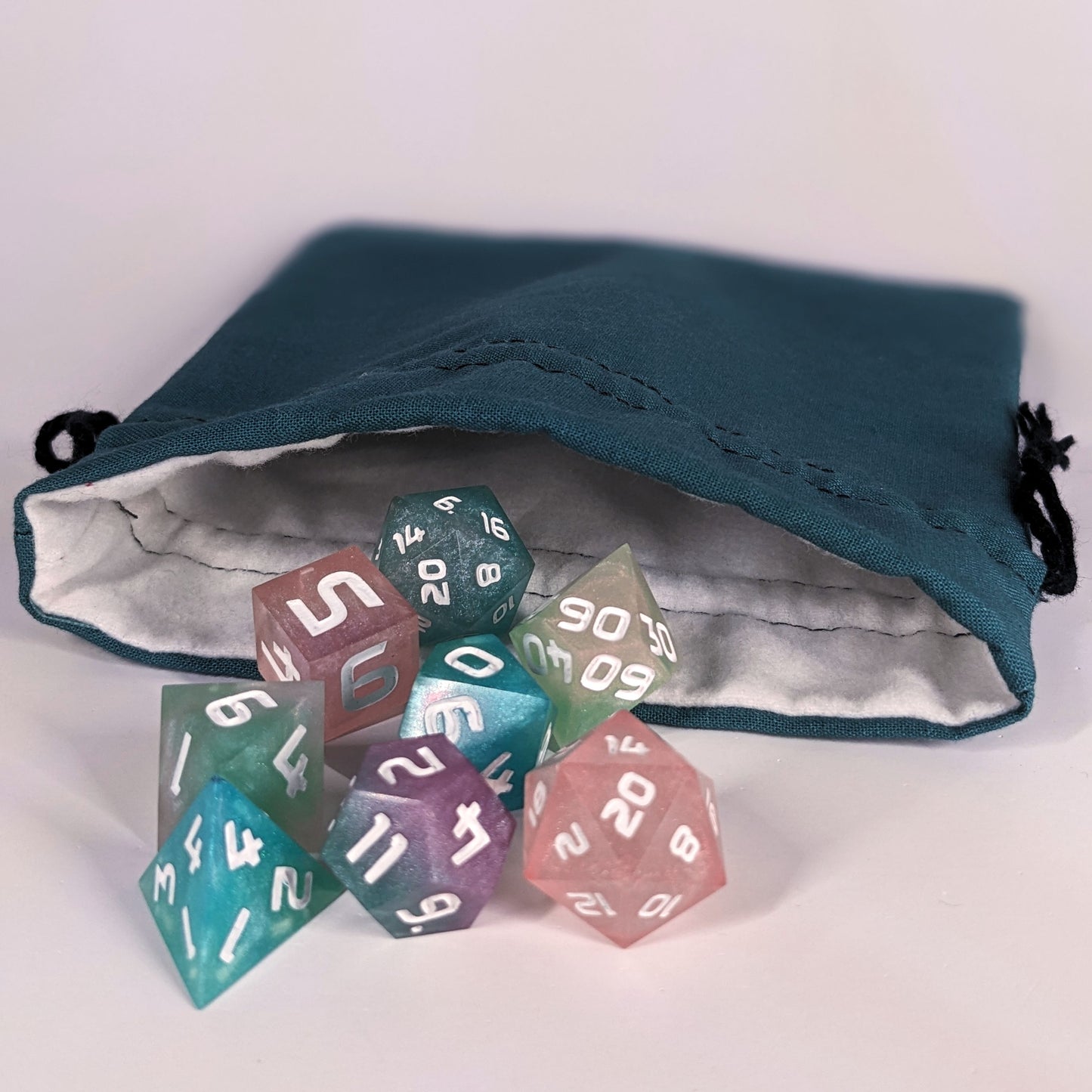 Colorful polyhedral d&d dice spill out of the open mouth of a teal, medium size dice bag. It shows the interior of the bag, which is a white flannel fabric, and the drawstrings are black.
