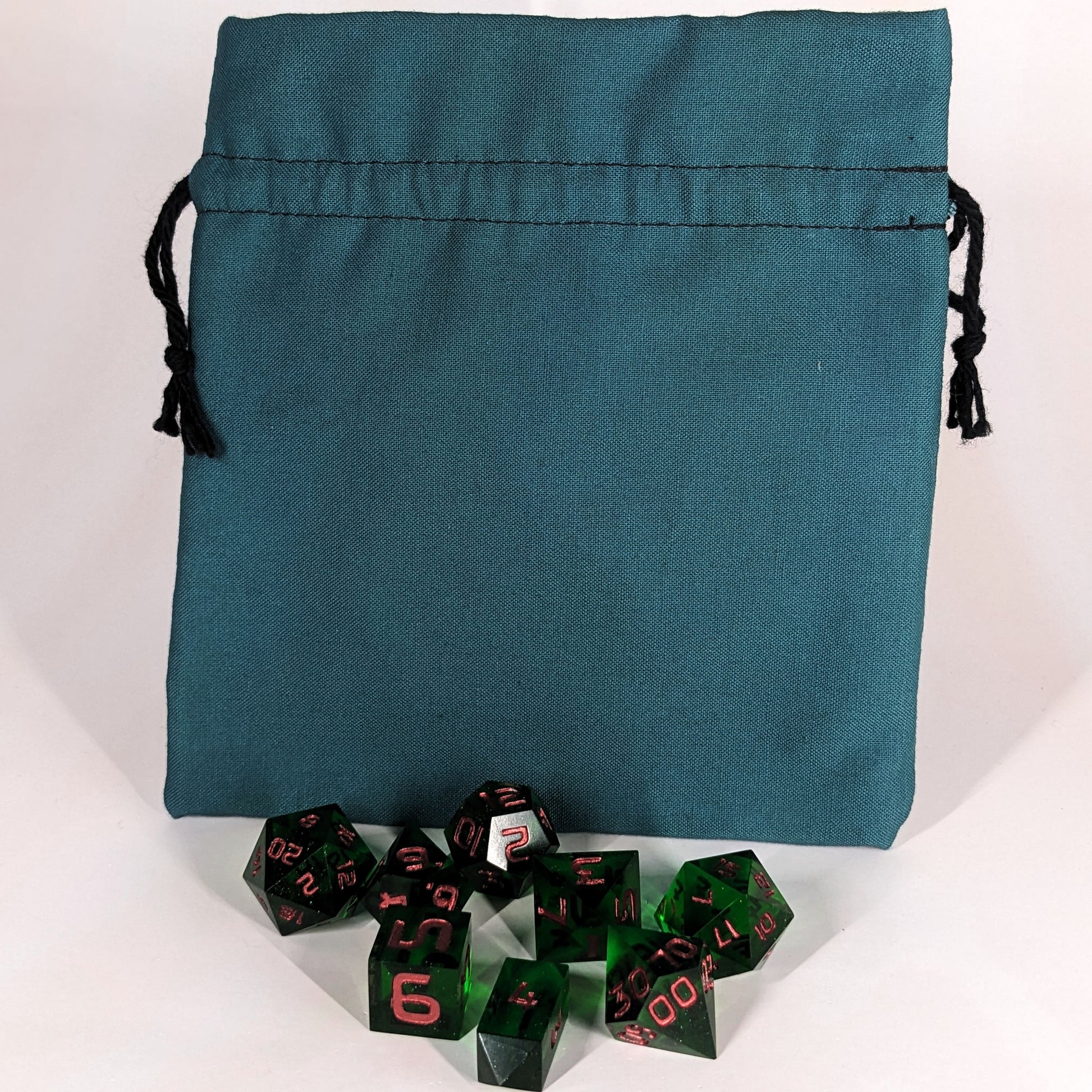 A set of 8 emerald green, handmade dnd dice sit in front of a teal, medium size dice bag that is sitting upright and not tied closed. The black drawstrings are visible on each side of the bag.