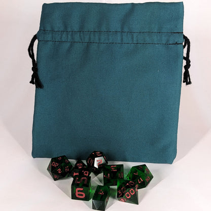 A set of 8 emerald green, handmade dnd dice sit in front of a teal, medium size dice bag that is sitting upright and not tied closed. The black drawstrings are visible on each side of the bag.