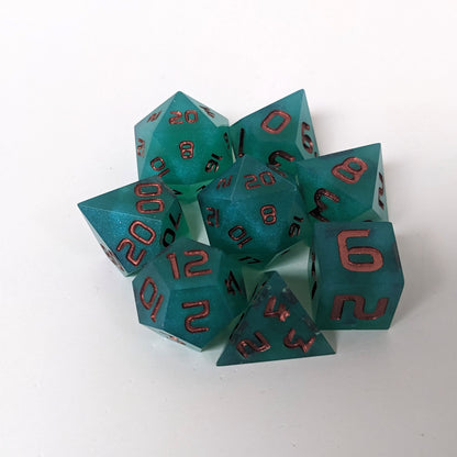 A complete set of 8 handmade artisan dice that are teal with a pearlescent shine with copper numbers. The DnD dice set is arranged in a circle and features 2 D20s, one of which is in the center surrounded by the other 7 dice on a white background.