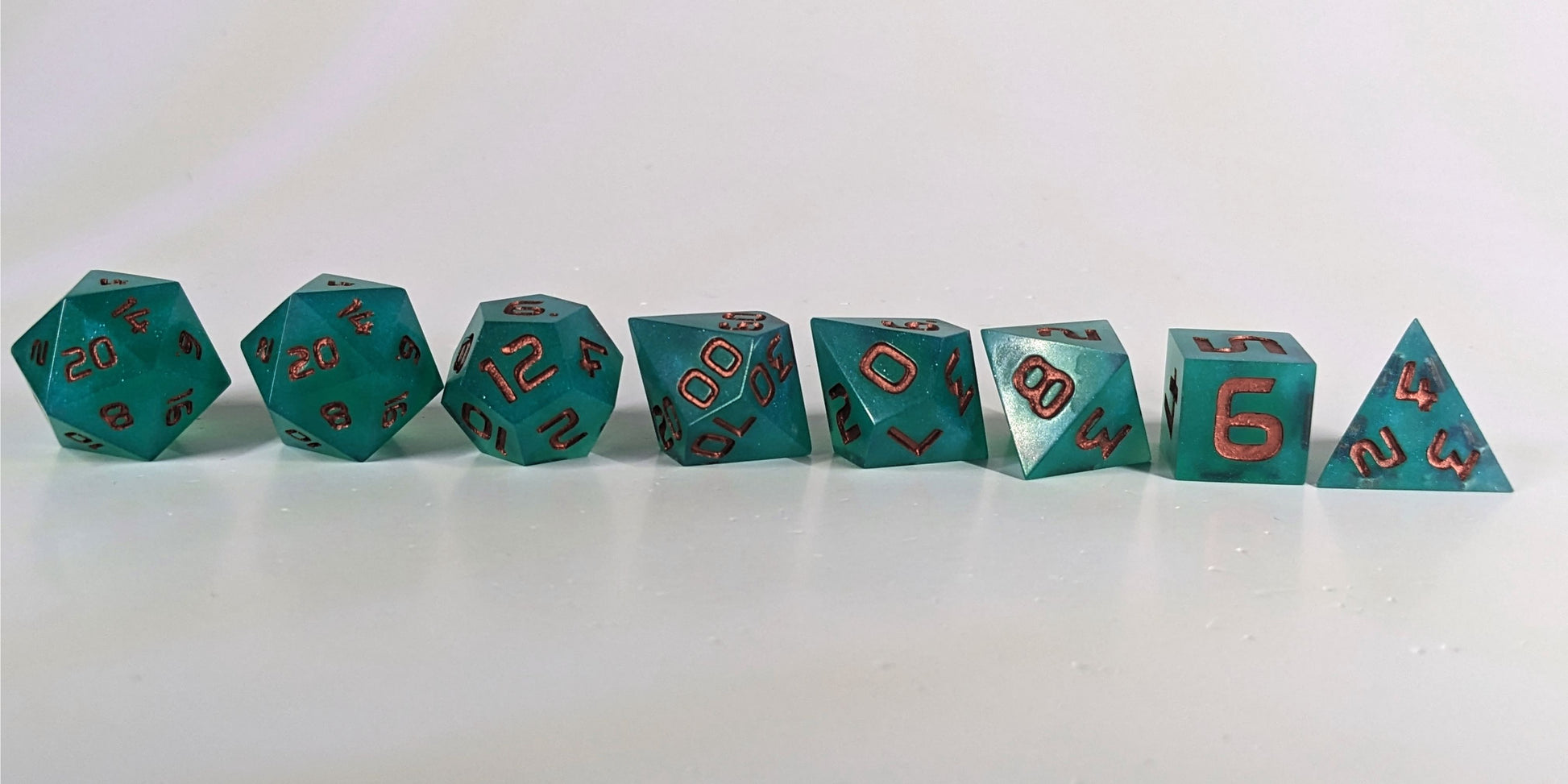 A straight on photo of a complete set of polyhedral dice that are teal with a pearlescent shine with copper numbers arranged in a straight line. They are lined up with the 2 d20s on the left going down to the d4 on the right side. The highest value number of each die is facing forward