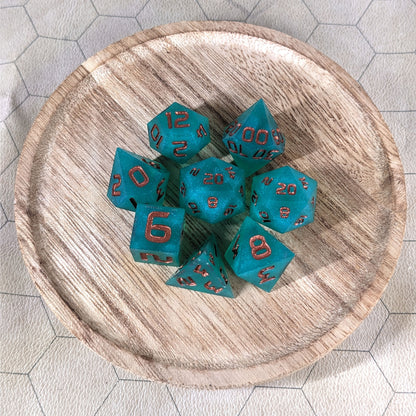 A complete set of 8 handmade artisan dice that are teal with a pearlescent shine and copper numbers. The DnD dice set is on a wooded circle and the dice are arranged in a circle and features 2 D20s, one of which is in the center surrounded by the other 7 dice.