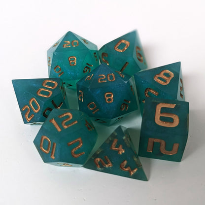 A complete set of 8 handmade artisan dice that are teal with a pearlescent shine with gold numbers. The DnD dice set is arranged in a circle and features 2 D20s, one of which is in the center surrounded by the other 7 dice on a white background.