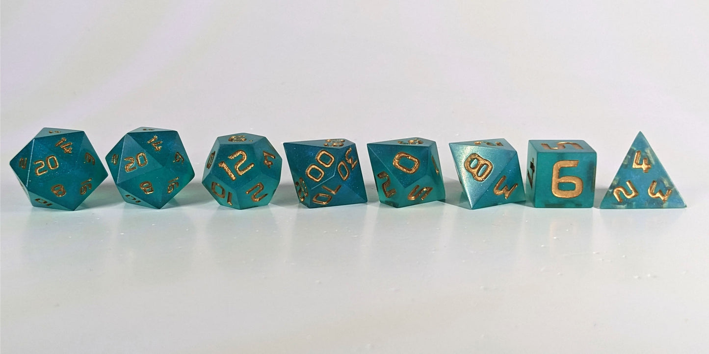 A straight on photo of a complete set of polyhedral dice that are teal with a pearlescent shine with gold numbers arranged in a straight line. They are lined up with the 2 d20s on the left going down to the d4 on the right side. The highest value number of each die is facing forward