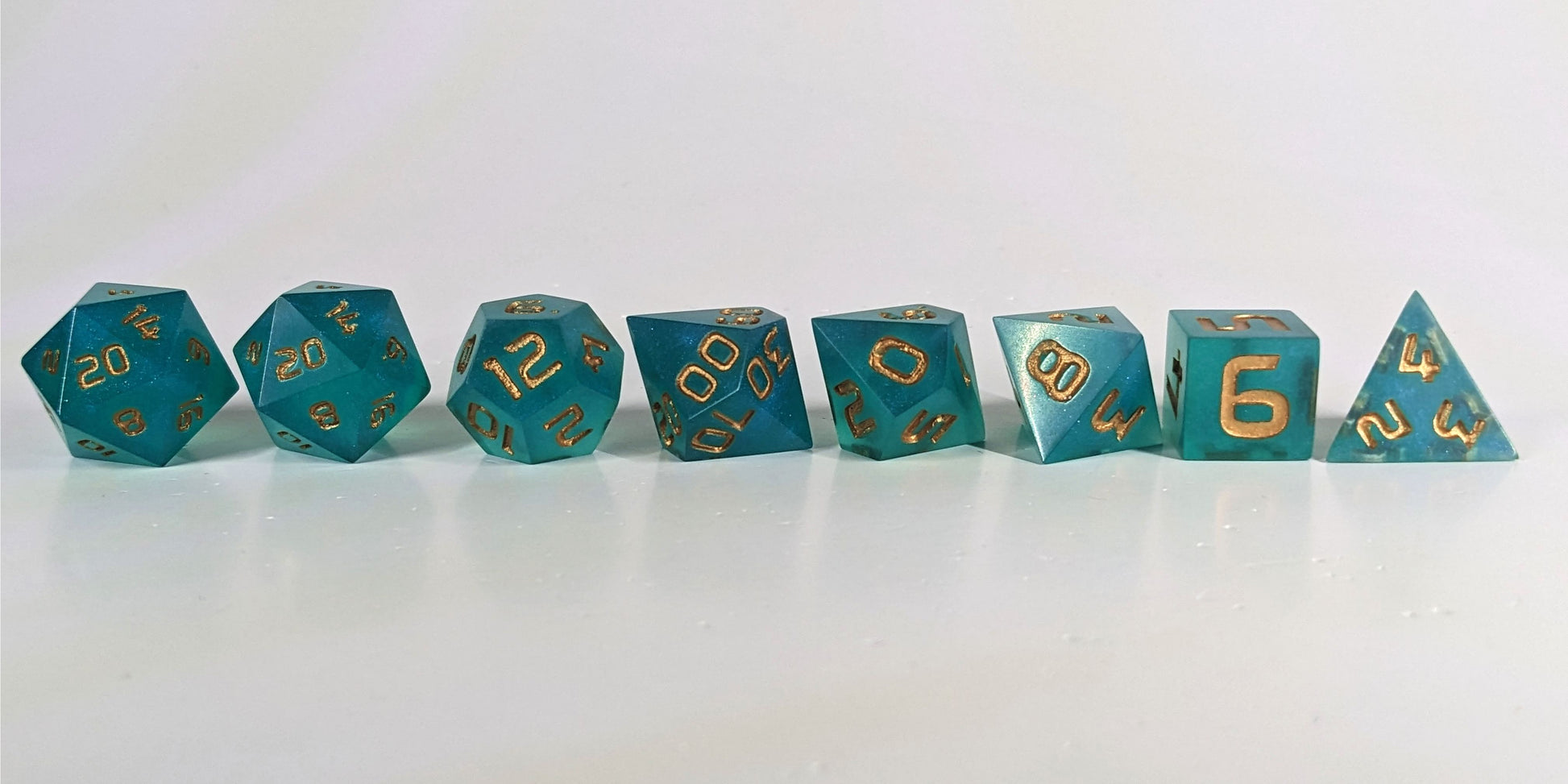 A straight on photo of a complete set of polyhedral dice that are teal with a pearlescent shine with gold numbers arranged in a straight line. They are lined up with the 2 d20s on the left going down to the d4 on the right side. The highest value number of each die is facing forward