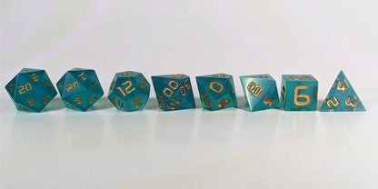 A straight on photo of a complete set of polyhedral dice that are teal with a pearlescent shine with gold numbers arranged in a straight line. They are lined up with the 2 d20s on the left going down to the d4 on the right side. The highest value number of each die is facing forward