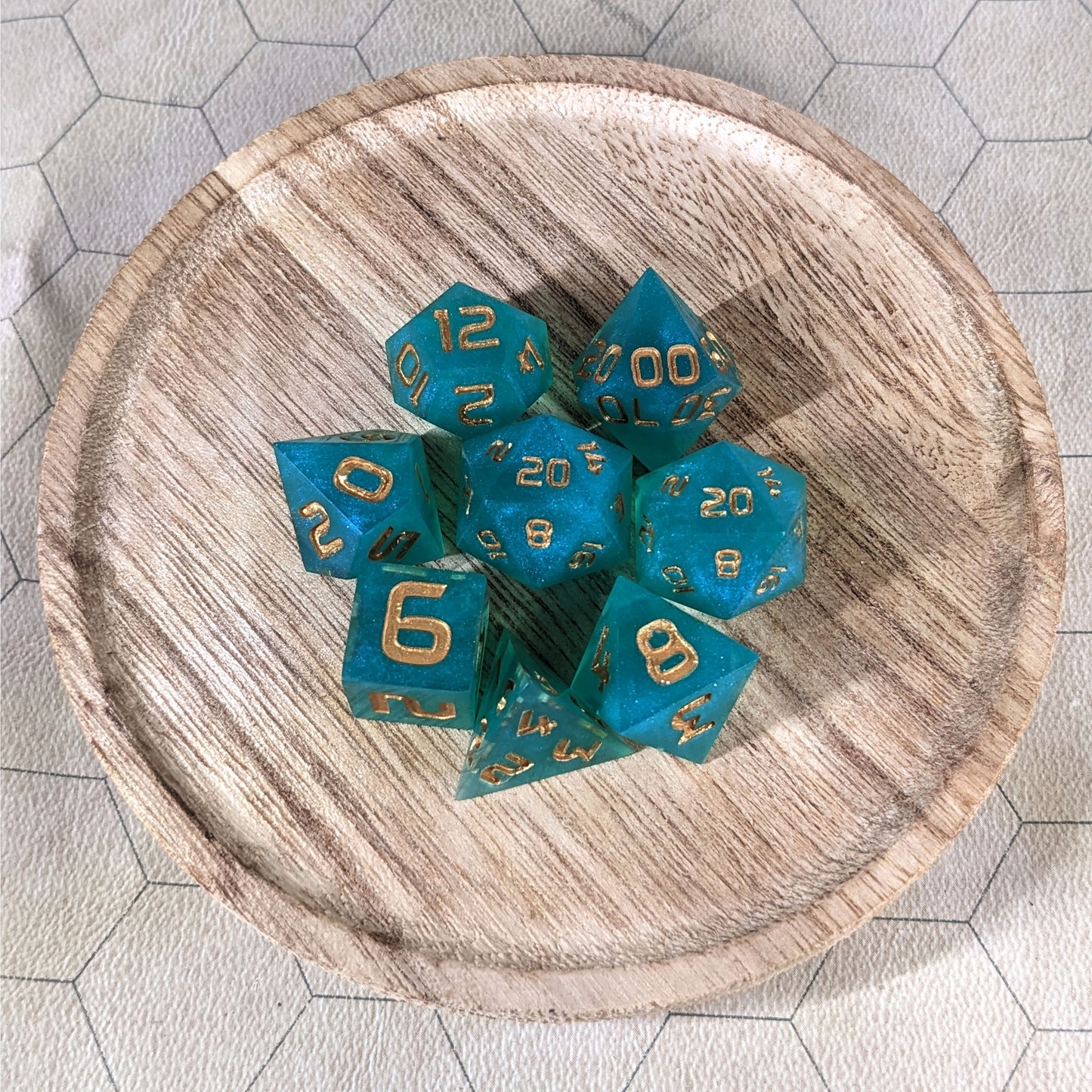 A complete set of 8 handmade artisan dice that are teal with a pearlescent shine and gold numbers. The DnD dice set is on a wooded circle and the dice are arranged in a circle and features 2 D20s, one of which is in the center surrounded by the other 7 dice.