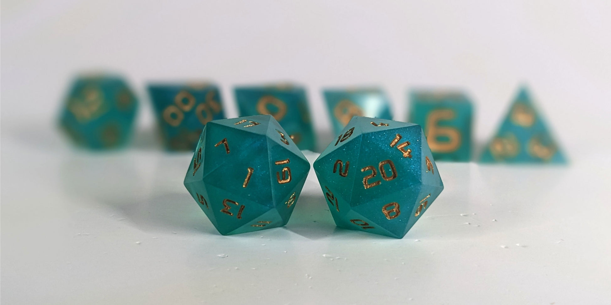 A straight on photo of a standard set of DnD dice that are teal with a pearlescent shine with gold numbers focusing on 2 d20s in the center. The d20 on the left has the 1 face centered and the d20 on the right has the 20 face centered. The other 6 dice are lined up in the background but are out of focus, while the d20s are in sharp focus.