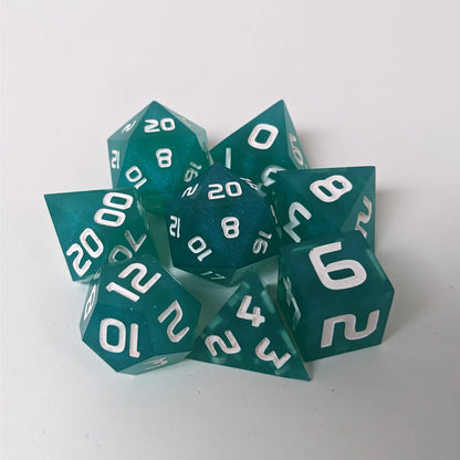 A complete set of 8 handmade artisan dice that are teal with a pearlescent shine with white numbers. The DnD dice set is arranged in a circle and features 2 D20s, one of which is in the center surrounded by the other 7 dice on a white background.