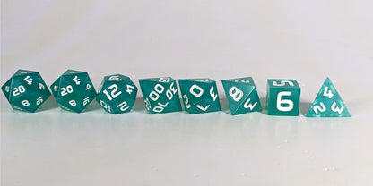 A straight on photo of a complete set of polyhedral dice that are teal with a pearlescent shine with white numbers arranged in a straight line. They are lined up with the 2 d20s on the left going down to the d4 on the right side. The highest value number of each die is facing forward