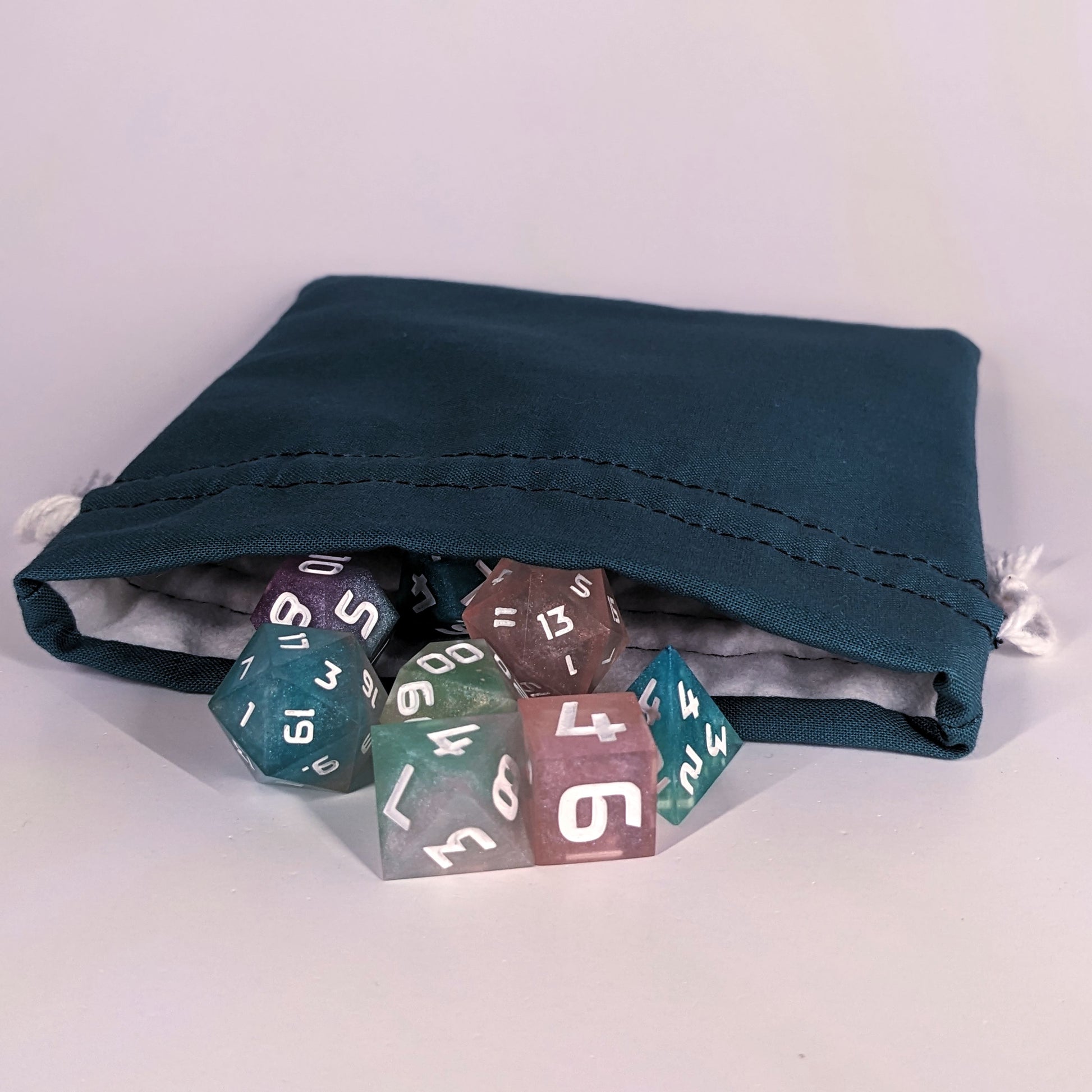 Colorful polyhedral d&d dice spill out of the open mouth of a teal, medium size dice bag. It shows the interior of the bag, which is a white flannel fabric, and the drawstrings are white.