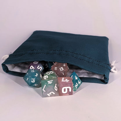 Colorful polyhedral d&d dice spill out of the open mouth of a teal, medium size dice bag. It shows the interior of the bag, which is a white flannel fabric, and the drawstrings are white.