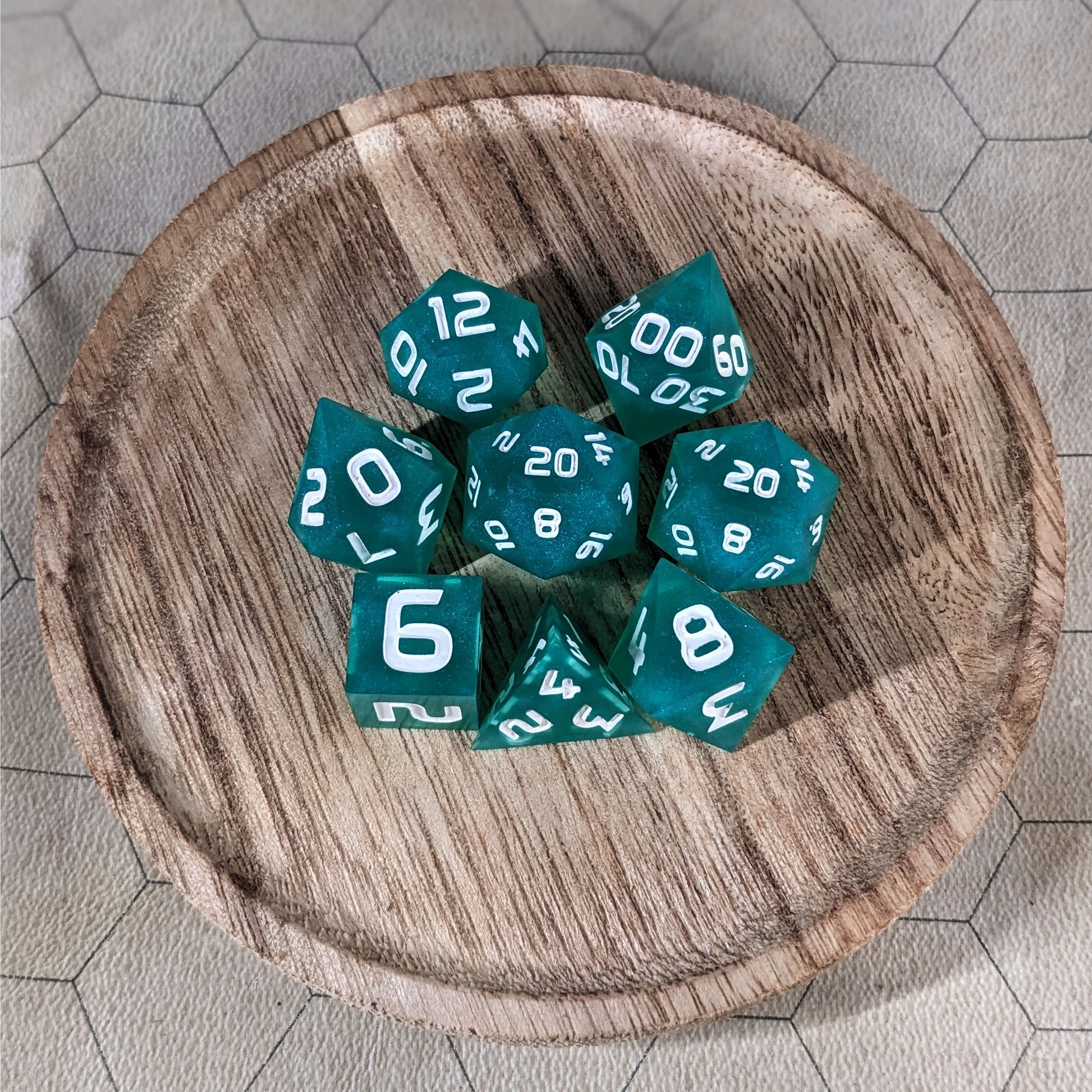 A complete set of 8 handmade artisan dice that are teal with a pearlescent shine and white numbers. The DnD dice set is on a wooded circle and the dice are arranged in a circle and features 2 D20s, one of which is in the center surrounded by the other 7 dice.