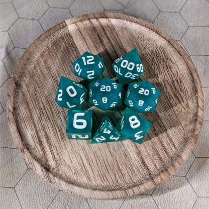 A complete set of 8 handmade artisan dice that are teal with a pearlescent shine and white numbers. The DnD dice set is on a wooded circle and the dice are arranged in a circle and features 2 D20s, one of which is in the center surrounded by the other 7 dice.