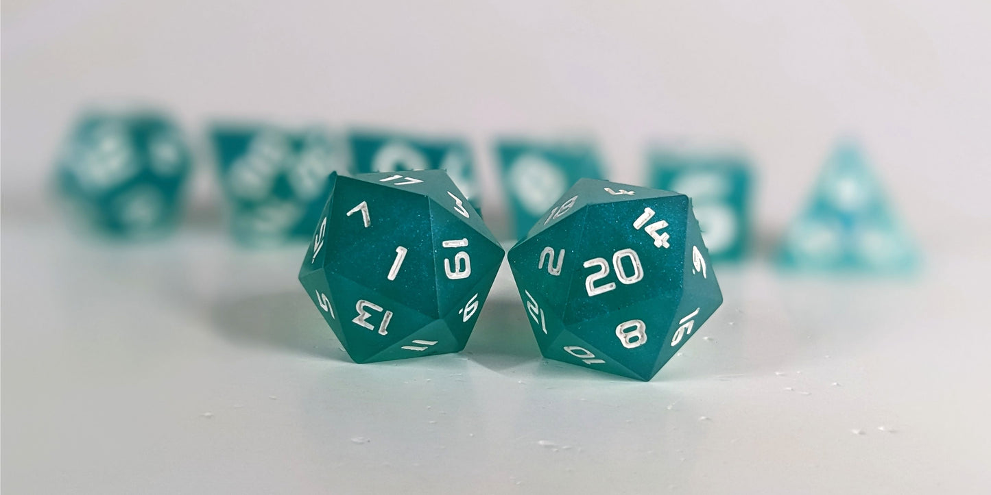 A straight on photo of a standard set of DnD dice that are teal with a pearlescent shine with white numbers focusing on 2 d20s in the center. The d20 on the left has the 1 face centered and the d20 on the right has the 20 face centered. The other 6 dice are lined up in the background but are out of focus, while the d20s are in sharp focus.