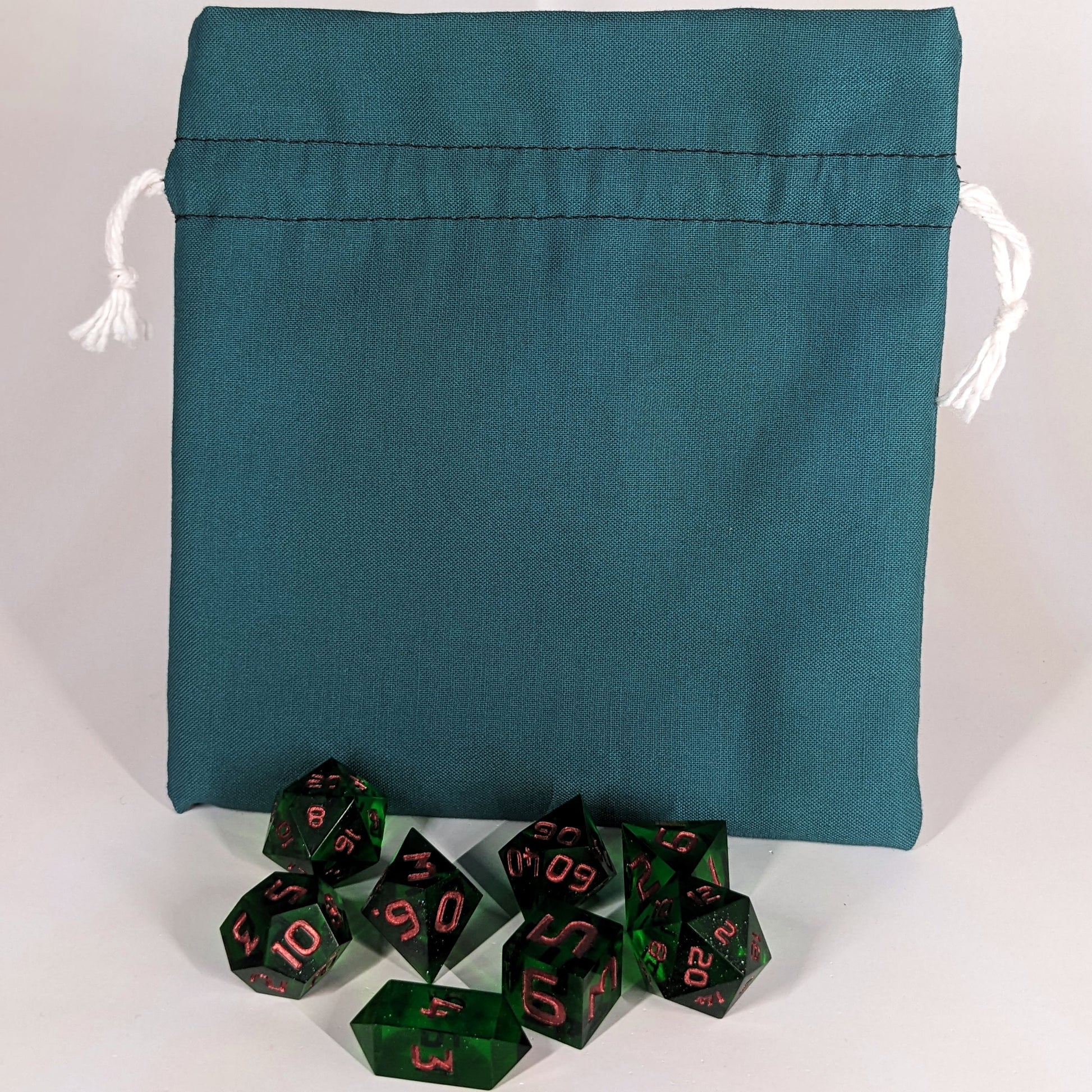 A set of 8 emerald green, handmade dnd dice sit in front of a teal, medium size dice bag that is sitting upright and not tied closed. The white drawstrings are visible on each side of the bag.