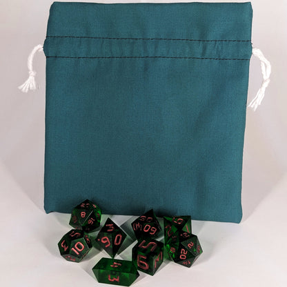 A set of 8 emerald green, handmade dnd dice sit in front of a teal, medium size dice bag that is sitting upright and not tied closed. The white drawstrings are visible on each side of the bag.