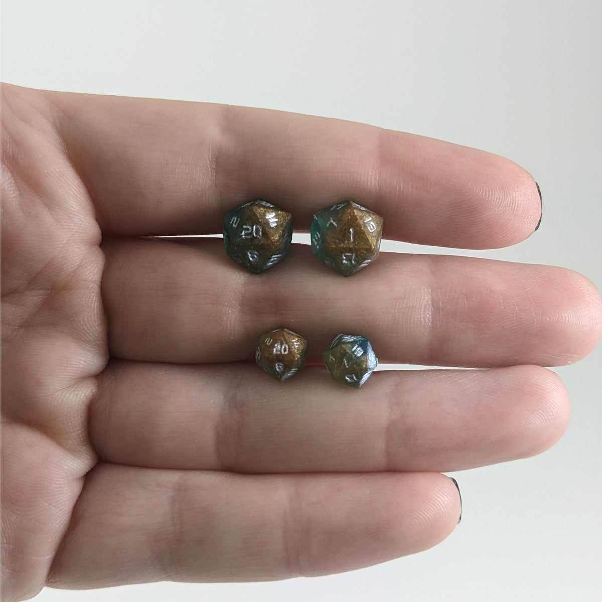 Two pairs of handmade d20 earrings with titanium hardware that are teal and copper swirls with silver numbers held between the fingers of a flat hand. The larger pair is between the pointer and middle finger, the smaller between the middle and ring finger. The earrings on the left of each pair centers the 20 face and the earrings on the right of each pair centers the 1 face.