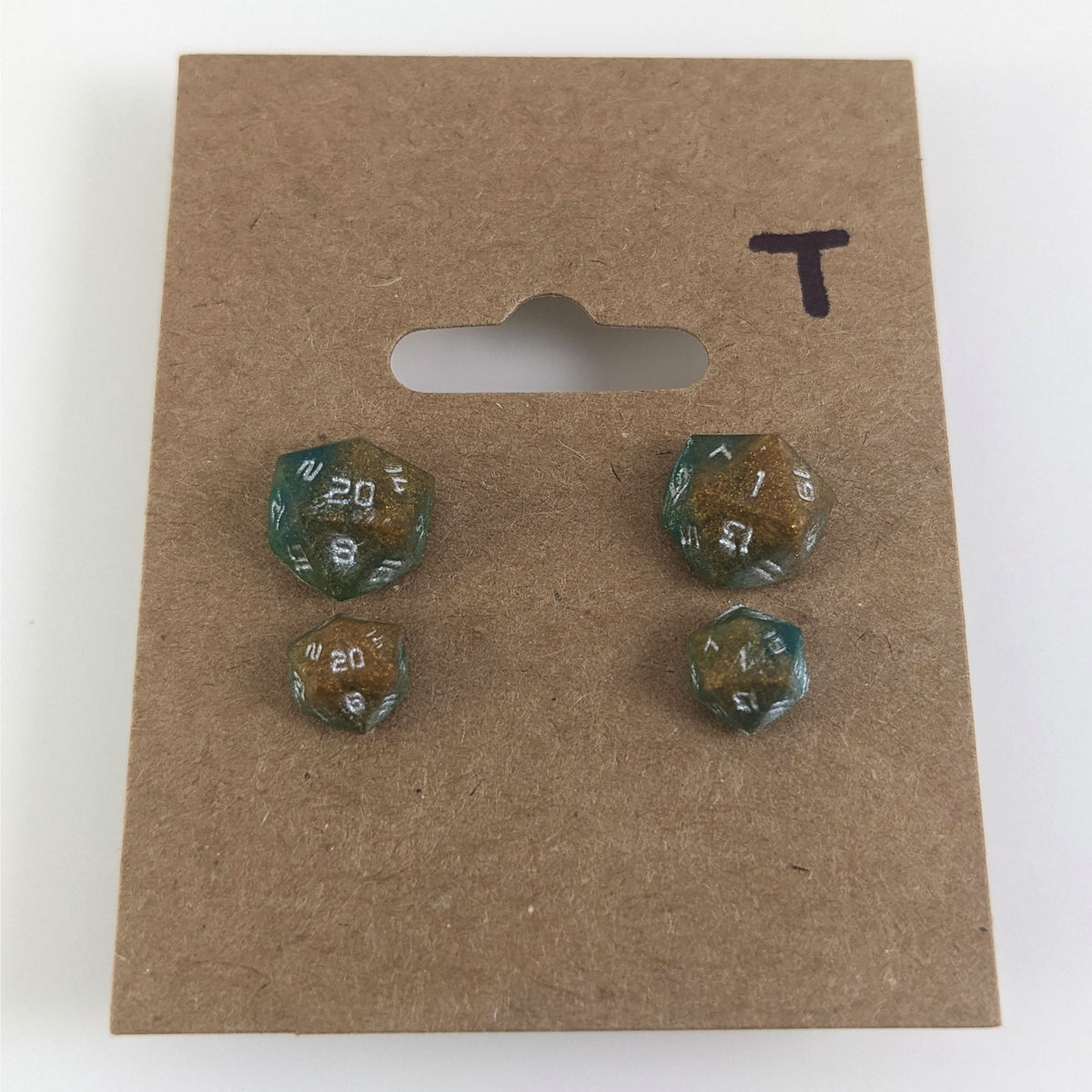 Two pairs of handmade d20 earrings with titanium hardware that are teal and copper swirls with silver numbers on a kraft brown earring card with a T in the top right corner. The earrings on the left of each pair centers the 20 face and the earrings on the right of each pair centers the 1 face.