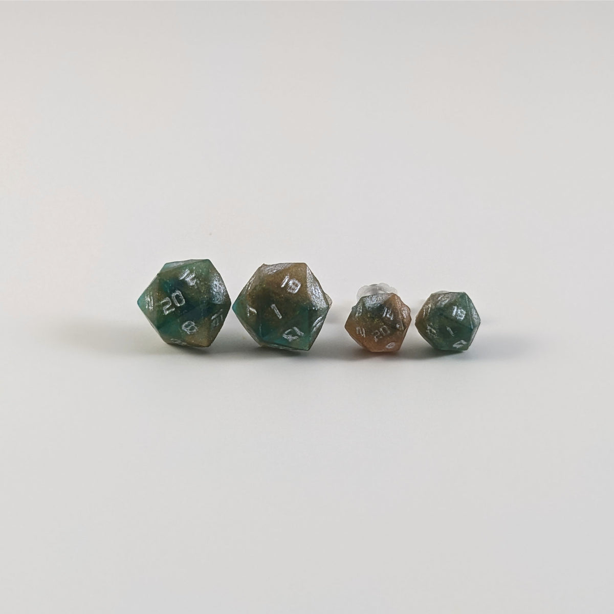 Two pairs of handmade d20 earrings with titanium hardware that are teal and copper swirls with silver numbers on a white background sit in a line with the larger pair on the left and the smaller pair on the right. The earrings on the left of each pair centers the 20 face and the earrings on the right of each pair centers the 1 face.