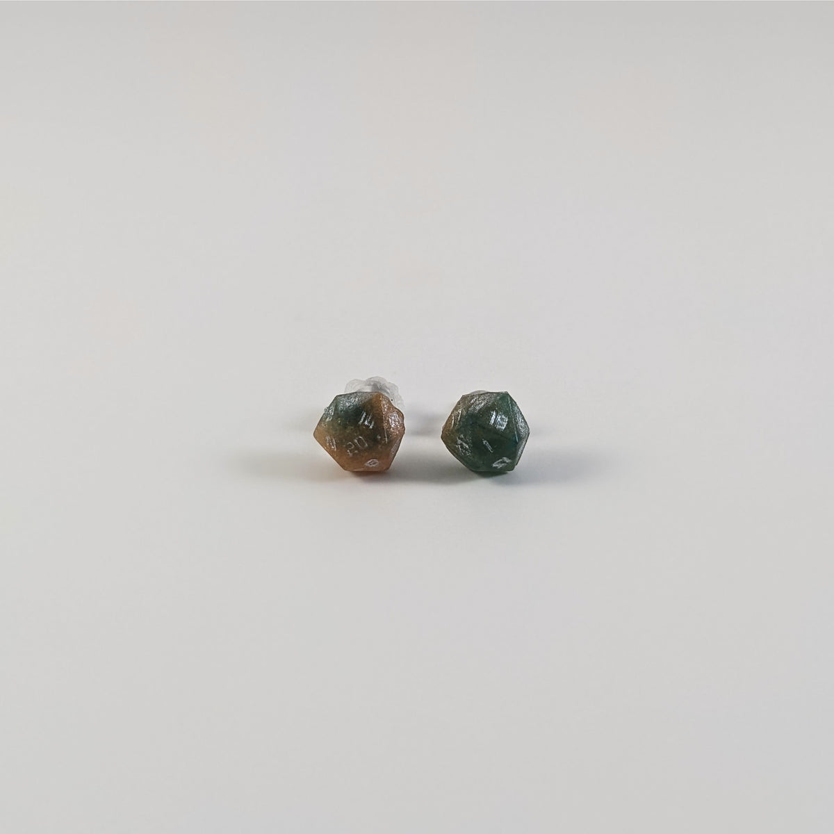 A single pair of the smaller size handmade d20 earrings with titanium hardware that are teal and copper swirls with silver numbers on a white background. The earring on the left centers the 20 face and the earring on the right centers the 1 face.